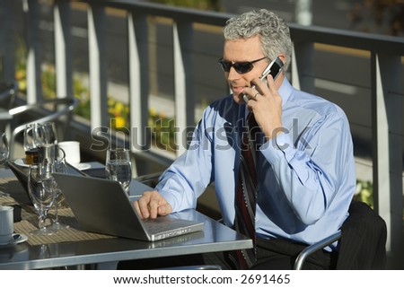 Prime adult Caucasian man in suit sitting at patio table outside looking at laptop and talking on cellphone.