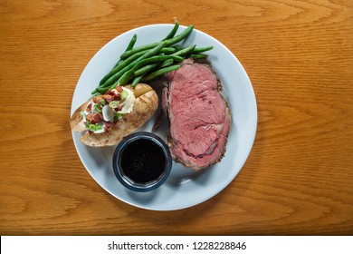 Prime Rib Meal Served On A Plate With A Loaded Potato And Green Beans With A Side Of A Jus