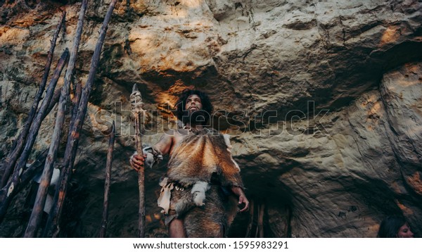 Primeval Caveman in Animal\
Skin and Fur Holds Stone Tipped Spear Comes out of His Cave into\
Prehistoric Forest Ready to Hunt. Neanderthal Going Hunting into\
the Jungle.