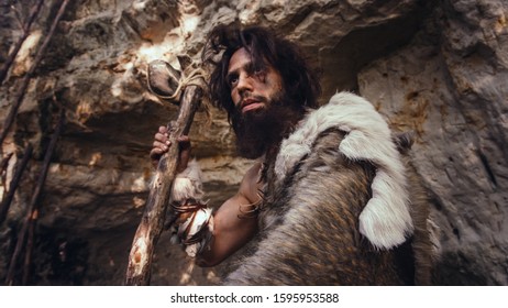 Primeval Caveman Wearing Animal Skin Holds Stone Hammer Looks Around Prehistoric Forest, Ready to Hunt Animal Prey. Neanderthal Going Hunting into the Jungle. Dramatic Low Angle Shot