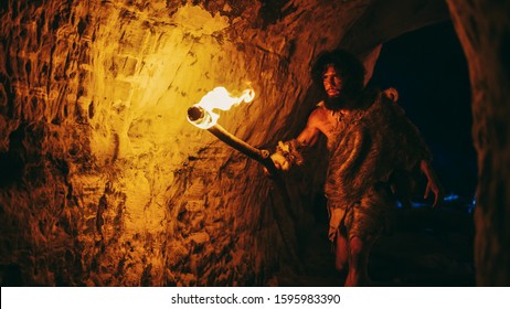 Primeval Caveman Wearing Animal Skin Exploring Cave At Night, Holding Torch with Fire Looking at Drawings on the Walls at Night. Neanderthal Searching Safe Place to Spend the Night