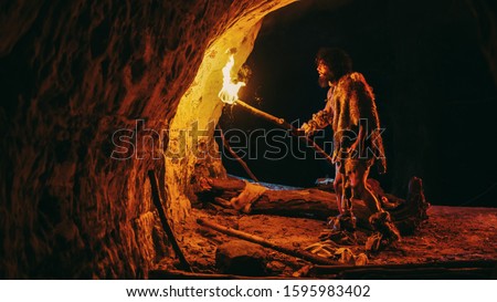 Primeval Caveman Wearing Animal Skin Exploring Cave At Night, Holding Torch with Fire Looking at Drawings on the Walls at Night. Cave Art with Petroglyphs, Rock Paintings. Side View
