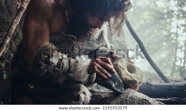 Primeval Caveman Wearing Animal\
Skin Hits Rock with Sharp Stone and Makes Primitive Tool for\
Hunting Animal Prey. Neanderthal Using Hand axe to Create first\
Wheel.
