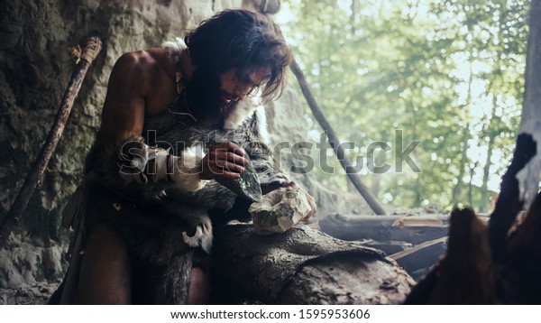 Primeval Caveman Wearing Animal Skin Hits\
Rock with Sharp Stone and Makes First Primitive Tool for Hunting\
Animal Prey or to Handle Hides. Neanderthal Using Handax. Dawn of\
Human Civilization