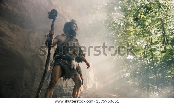 Primeval Caveman Wearing Animal Skin Holds Stone\
Hammer Stands Near Cave and Looks Around Prehistoric Landscape,\
Ready to Hunt Animal Prey. Neanderthal Going Hunting into Jungle.\
Low Angle Shot