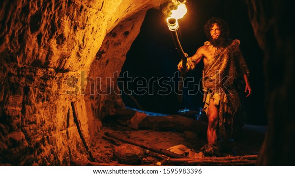 Primeval Caveman\
Wearing Animal Skin Exploring Cave At Night, Holding Torch with\
Fire Looking at Drawings on the Walls at Night. Neanderthal\
Searching Safe Place to Spend the\
Night