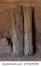 Primitive grass and juncus rush mats leaning against adobe brick wall inside dirt floored hut