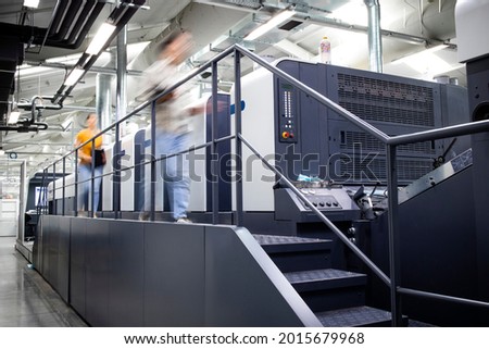 Printing factory interior with modern offset printing machine and busy people working around.