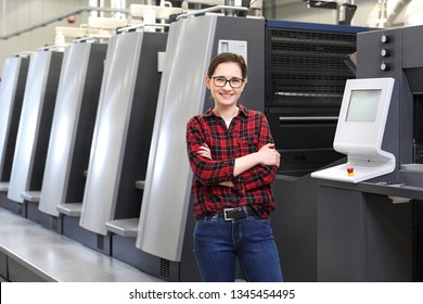 Printing house, bookbinding.
A smiling woman, an employee of the printing house stands in the production hall on the background of modern printing machines.
