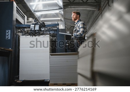 Printing house worker operating a large industrial printing machine, feeding paper sheets for commercial printing production