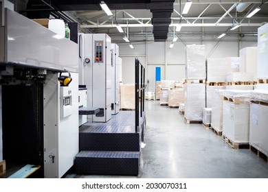 Printing shop or factory interior with modern offset paper machine and piles of sheets in background.