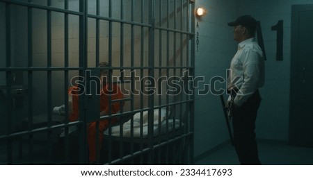 Prison officer walks the corridor, gives phone to female prisoner in orange uniform through metal bars in prison cell. Women serve imprisonment term in jail. Detention center or correctional facility.