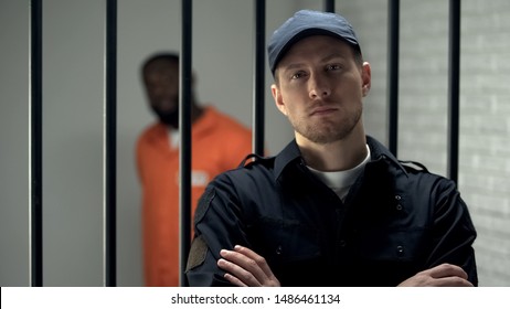 Prison Warden Looking To Camera Standing Near Cell With Imprisoned Afro-american