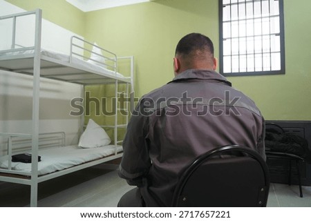 The prisoner is sitting in front of the bed in the cell. The colony.