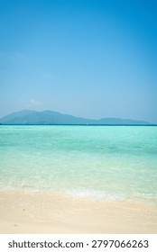 A pristine beach scene showcasing soft golden sand meeting crystal-clear turquoise waters. The horizon features lush green mountains, creating a tranquil and inviting tropical escape. Perfect for travel, tourism, and vacation marketing. Ideal for beach resort promotions, travel brochures, and relaxation-themed content. Sunlit and vibrant, this image captures the essence