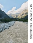 A pristine mountain river flows through a wide valley, framed by rugged peaks and lush forest slopes. Sunlight sparkles on the water, creating a tranquil alpine scene.