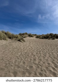 Pristine seaside landscape featuring rolling sand dunes along an empty beach under a clear vibrant blue sky, with hardy green shrubs dotting the sandy hillsides and adding texture and life to the natural scene. This peaceful image captures the quiet beauty of an untouched shoreline, where wind-sculpted dunes rise gently above the shore and native vegetation thrives in the salty air. With no people in sight, the photograph evokes a profound sense of solitude, freedom, and the meditative quality of wild seaside environments. The composition balances natural elements beautifully, with the soft curves of the dunes leading the eye across the frame while the green shrubs provide organic contrast against the pale golden sand and bright open sky above. The fresh color palette of warm sandy tones, vibrant greens, and crisp clear blues creates an uplifting, summery atmosphere perfect for evoking exploration, escape, and reverence for the natural world. This idyllic beachside scene captures the essence of a perfect day at the shore, making it ideal for travel publications, tourism marketing, beach resort and hotel branding, wellness and mindfulness content, environmental and conservation storytelling, real estate listings for coastal properties, lifestyle blogs, editorial features about hidden beaches and protected shorelines, eco-tourism promotion, surf and outdoor lifestyle brands, and design projects requiring vibrant atmospheric imagery. The dunes add texture, depth, and a sense of place, while the green shrubs suggest the resilience of life in challenging seaside ecosystems and the rich biodiversity of these often-overlooked habitats. With its combination of dramatic natural scenery, peaceful empty terrain, and complete absence of human presence, this photograph captures a quintessentially seaside moment where land meets sky in a celebration of unspoiled beauty. Perfect for website backgrounds, social media content, print advertising, travel brochures, postcards, wall art, book covers, calendars, and editorial layouts.