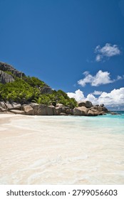 Pristine white sand beach with dramatic granite boulders and turquoise ocean waves on Big Sister Island, Seychelles under a clear blue sky.