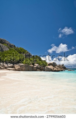 Pristine white sand beach with dramatic granite boulders and turquoise ocean waves on Big Sister Island, Seychelles under a clear blue sky.