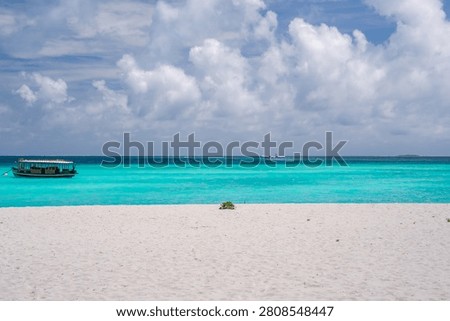 Pristine white sand meets shallow turquoise water under cumulus clouds. A covered boat floats left, a distant skiff appears offshore at a Maldives luxury resort.