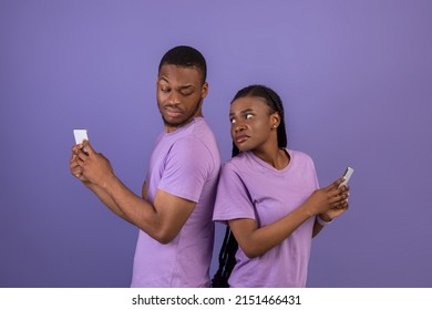 Privacy, Distrust, Infidelity Concept. Curious Angry Black Man Trying To Look At His Girlfriend's Smartphone, Confused Woman Hiding Screen Of Her Gadget, Couple Standing Over Purple Studio Background