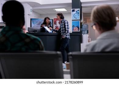 Private Clinic Receptionist Giving Assistance To Asian Patient To Complete Hospital Admission Form At Front Desk. Selective Focus On Young Man Talking With Woman Holding Clipboard With Medical Data.