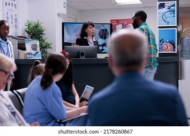 Private Clinic Receptionist Giving Informations To African American Patient Registering For Doctor Appointment At Front Desk. Man In Hospital Waiting Room Asking For Healthcare Details In Busy Clinic.