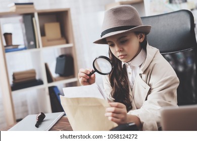 Private detective agency. Little girl in cloak and hat is sitting at desk looking at photos with magnifying glass. - Shutterstock ID 1058124272
