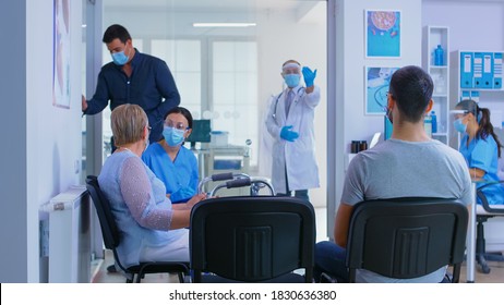 Private Hospital Reception Area During Global Pandemic. Doctor With Visor Against Coronavirus Inviting Man In His Office For Examination. Disabled Senior Woman With Walking Frame