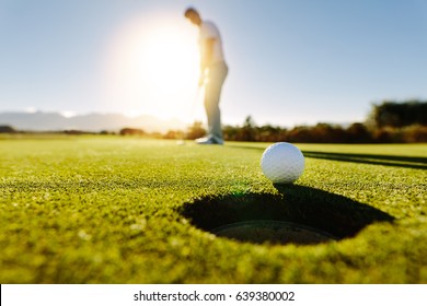 Pro golfer putting golf ball in to the hole. Golf ball by the hole with player in background on a sunny day.