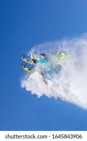 A Pro Snowmobiler Makes A Jump With A Big Blast Of Snow And Epic Falls. Background Of Blue Sky Leaving A Trail Of Splashes Of White Snow. Bright Snow Bike And Suit. Snowmobile