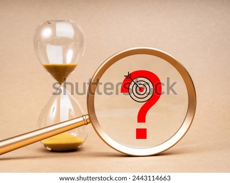 Problem analysis and solving, time to making right decision concept. Red question mark icon with target symbol in big magnifying glass lens near golden hourglass on light brown background.