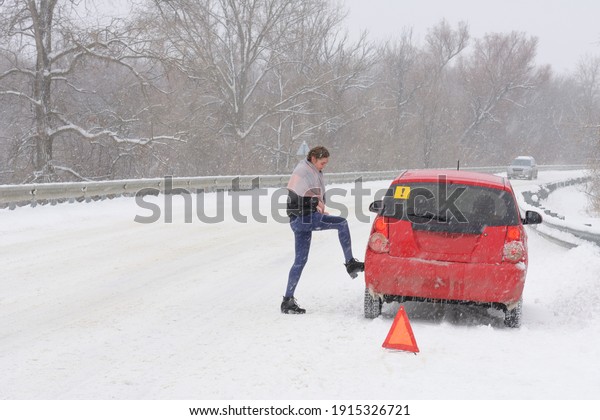 Problem with car on  snowy road in blizzard.\
Woman novice driver touches tire with  foot. Triangular accident\
warning sign stands on \
roadway.
