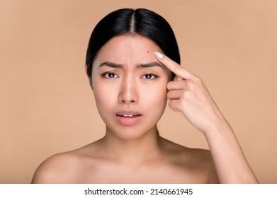 Problem skin concept. Upset asian girl with nude shoulders, with problem skin, pointing finger at pimple on forehead while standing over isolated beige background, sadly looks at camera