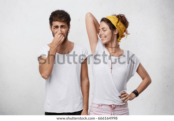 Problems with body odor. Disgusted male pinching\
his nose feeling bad smell or stink coming out from attractive\
smiling girl, who is raising her arm, showing wet t-shirt because\
of armpit sweat