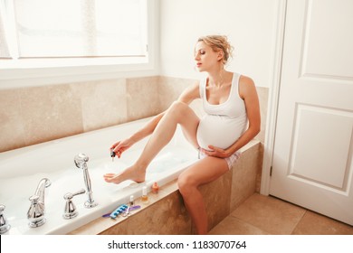 Problems And  Challenges During Pregnancy. Pregnant Blonde Caucasian Woman Painting Polishing Finger Nails On Legs Toes In Bathroom. Expecting Mom Doing Cosmetological Procedures.