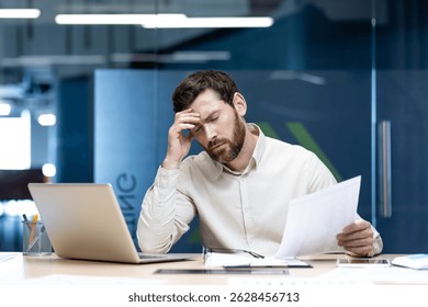 Problems with documents and data. A young man holds papers in his hands, sits at his desk and holds his head in frustration.