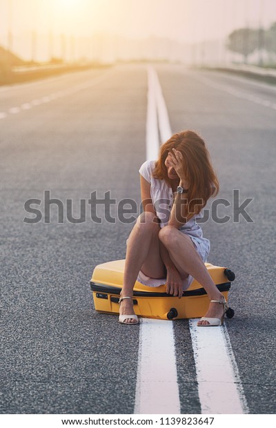 problems in the journey.\
the girl is sitting on a yellow suitcase on the dividing strip on\
the road.\
