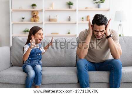 Problems Of Parenthood Concept. Portrait of angry naughty girl screaming loud and shouting at her tired father. Upset man covering ears, doesn't want to hear hysteria. Quarrel with shocked parent