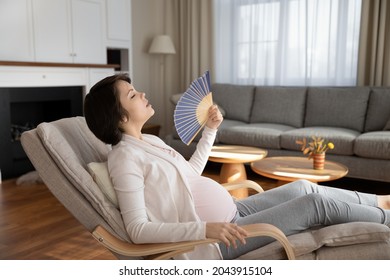 Problems of pregnancy. Exhausted pregnant female sit on chair cool herself using hand fan feel too hot in living room without air conditioner. Expectant mother suffer of heat indoors need ventilation
