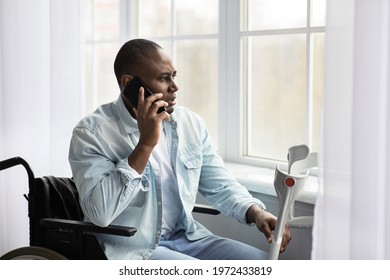 Problems At Work, Frustration And Bad News. Anxious, Sad, Tense Adult African American Disabled Man In Wheelchair With Crutches Talking On Phone And Looking At Window In House Or Clinic Interior