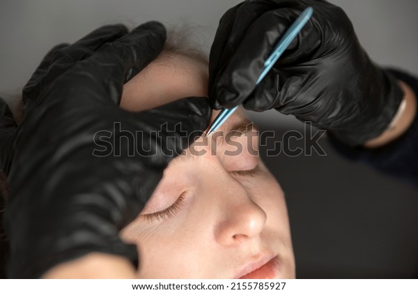 Procedure\
of eyebrow correction, young woman in beauty salon. Cosmetologist\
in black gloves plucks eyebrows with\
tweezers