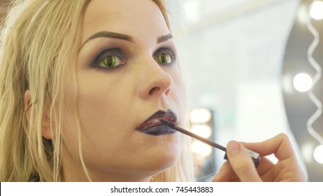 Process applying halloween makeup on face the young beautiful blonde woman contact lenses as cat eyes. Artist applying lipstick.