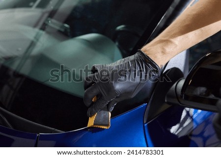 The process of applying a nano-ceramic coating on the white car's hood by a male worker in blue gloves with a sponge and chemical composition to protect the paint in detailing workshop. Auto service.