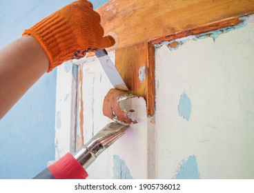 The Process Of Cleaning An Old Wooden Door From Paint Layers. Removal Of The Coating With A Hair Dryer And A Spatula. Worker Hands In Orange Gloves With A Tool. Household Renovation.