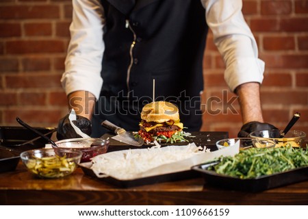 Process of cooking burgers. Cropeed view of chef hands in black gloves with prepared cheeseburger, variety of fillings and ingridients on wooden desk. Catering and Good Quality Fastfood concept