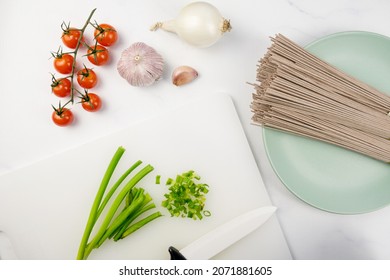 The Process Of Cooking Soba - Buckwheat Japanese Noodles. On A Cutting Board, Chopped Green Onions, Uncooked Soba, Garlic, White Onions, Cherry Tomatoes. Vegetarian Dish, Healthy Balanced Diet.