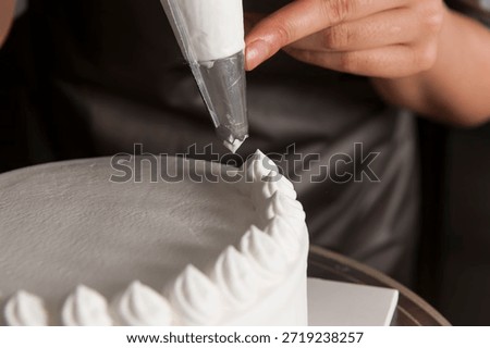 The process of decorating a cake: Pastry chef using a piping bag to create white whipped cream swirls on a fresh sponge cake. 