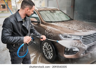 Process Of Detailing Car Cleaning. Guy In Casual Clothes Spraying Water Under High Pressure On His Automobile, Using Water Gun. Non-contact Cleaning Of Auto Body At Car Wash.