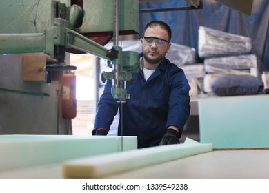 Process of furniture production. A young man in a dark coat cut the foam for the sofa on a cutting machine. Behind him there are a lot of finished sofas. He is wearing a safety glasses and gloves. - Powered by Shutterstock